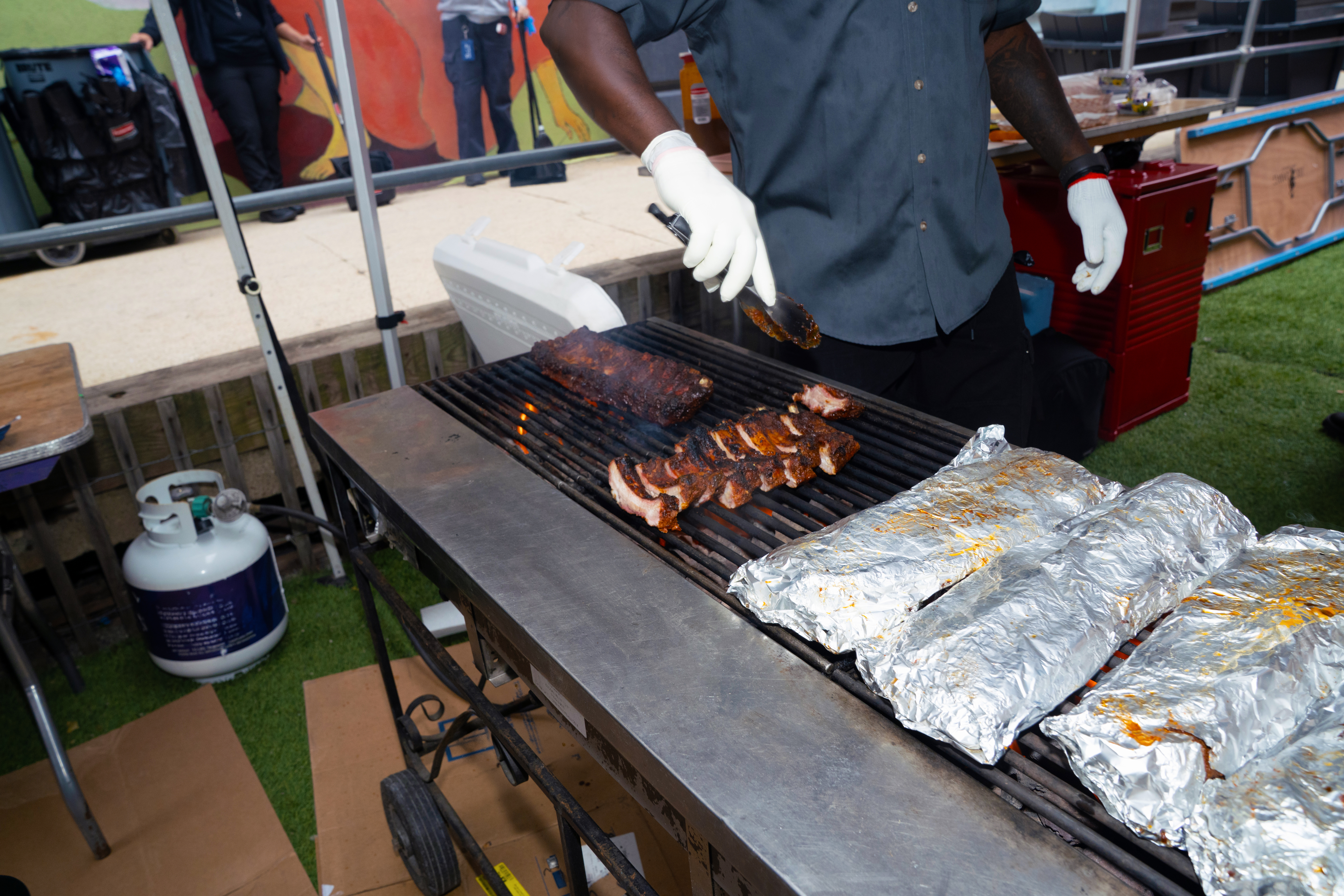 A street food vendor wearing black gloves expertly slices freshly cooked barbecue ribs on a wooden cutting board at an outdoor market event.